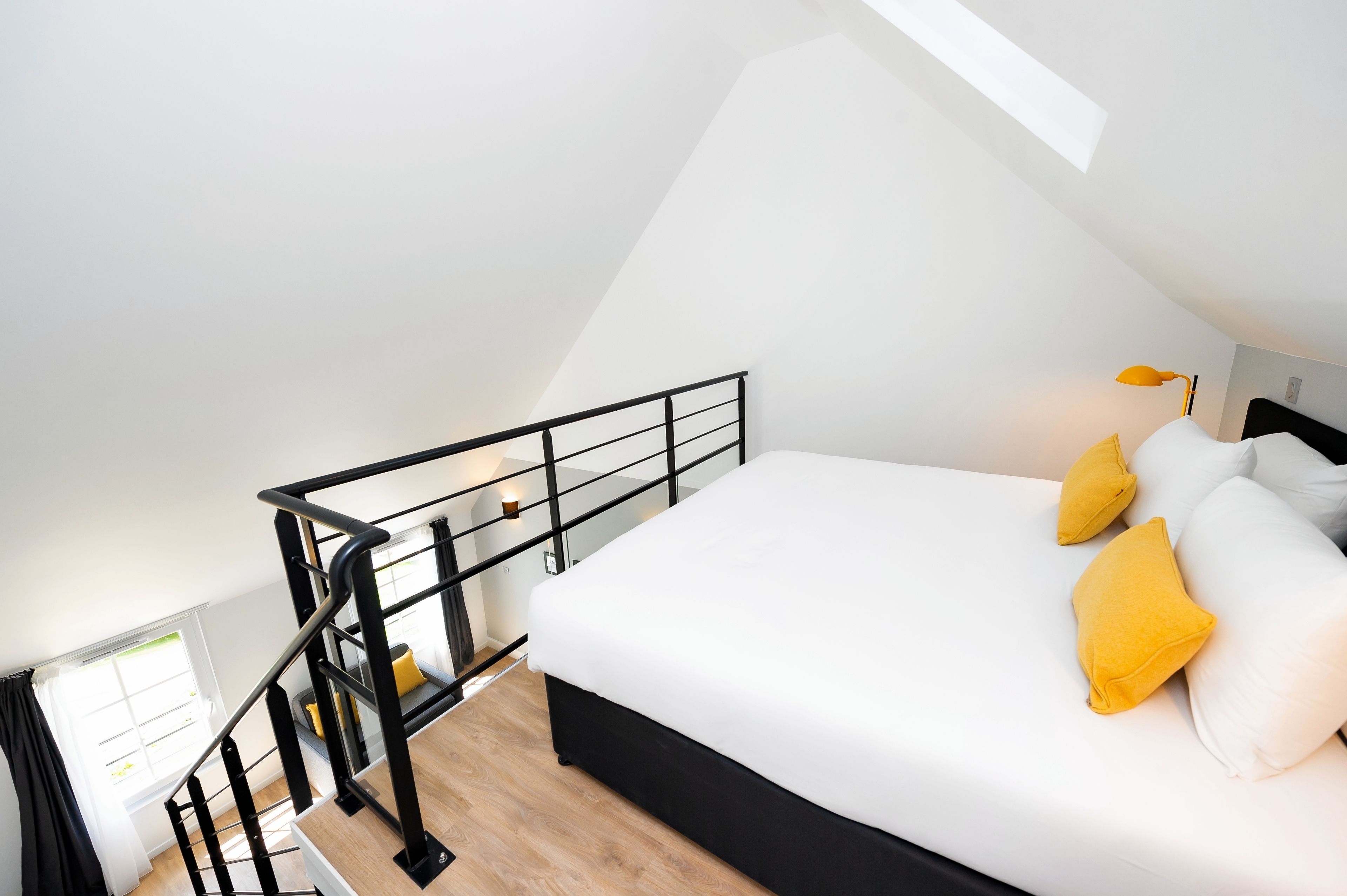 A bedroom with a white bed, black headboard, and yellow pillows. There is a black railing and a window with curtains.
