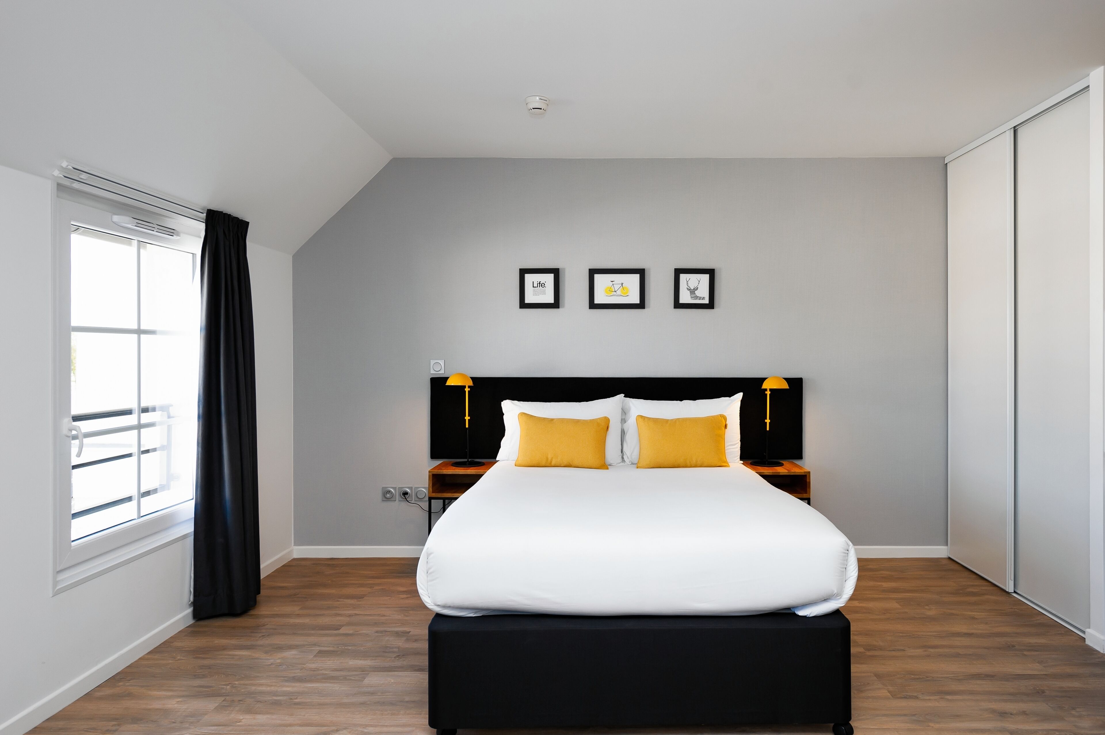 Un dormitorio moderno con una cama grande, una ventana con cortinas, un cabecero con dos lámparas y tres cuadros en la pared.