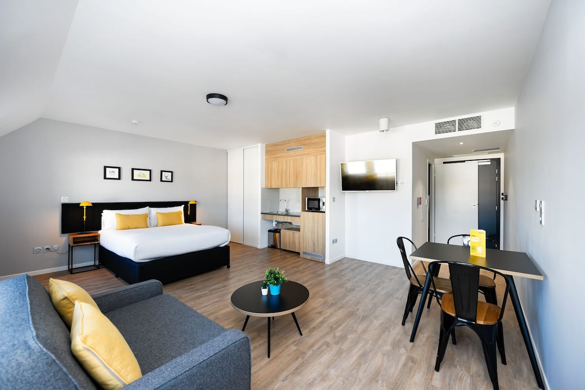 A modern hotel room with a bed, a sofa, a dining table, and a kitchen area.