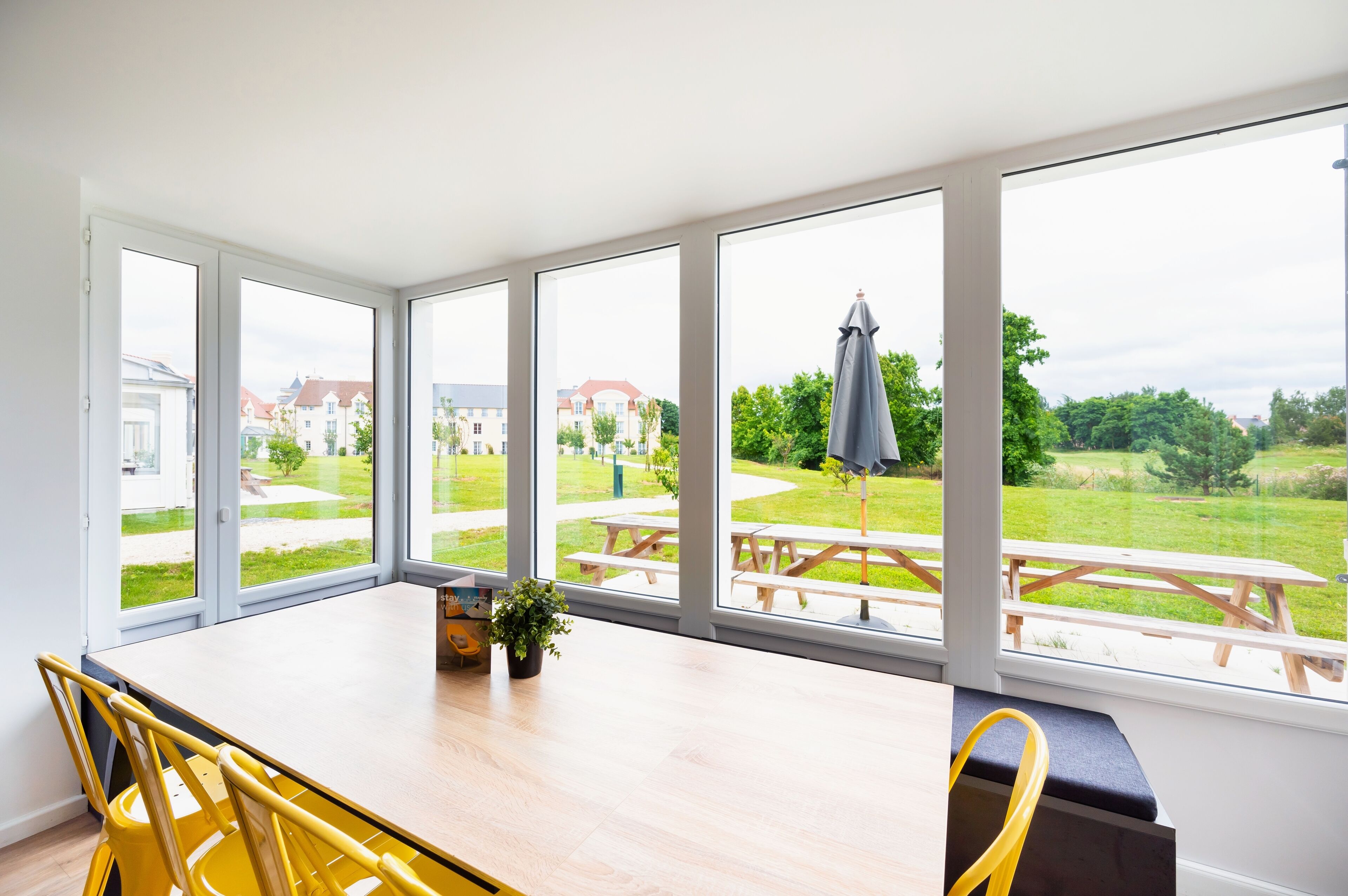 Una habitación luminosa con ventanas amplias, una mesa de madera, sillas amarillas y un banco.