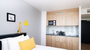 A compact room with a bed, a bedside table with a lamp, a small kitchenette with a microwave, kettle, and coffee maker.
