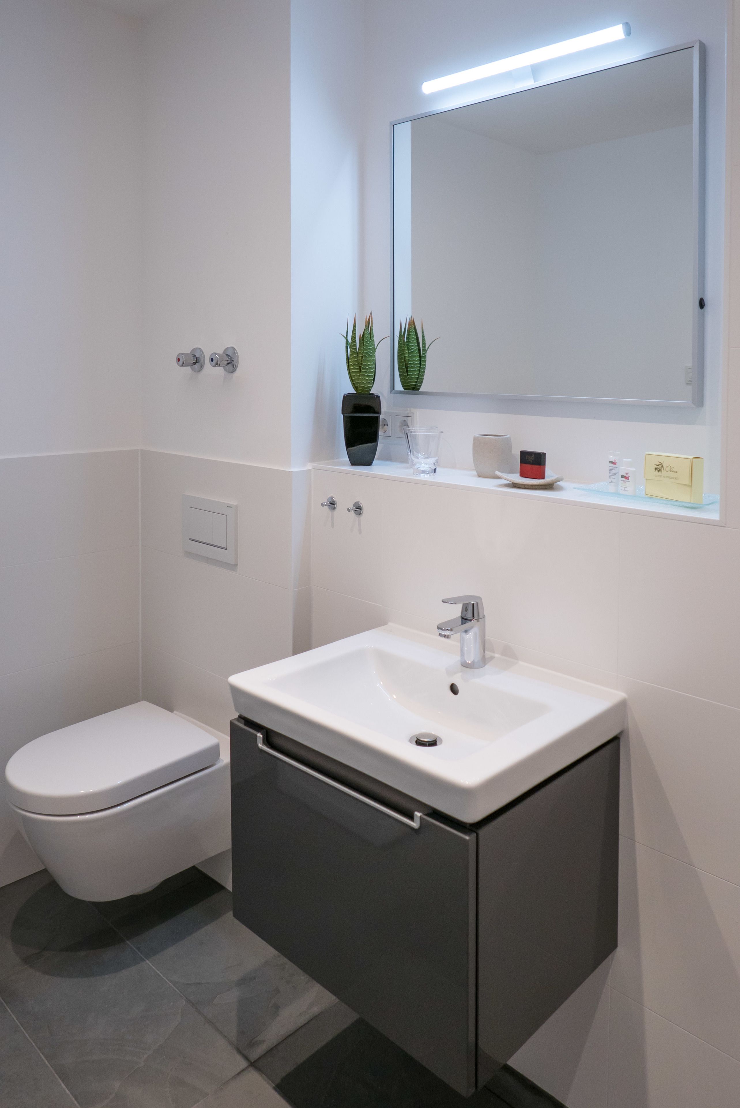 A modern bathroom with a white toilet, a white sink with a mirror above, and a shelf with toiletries.