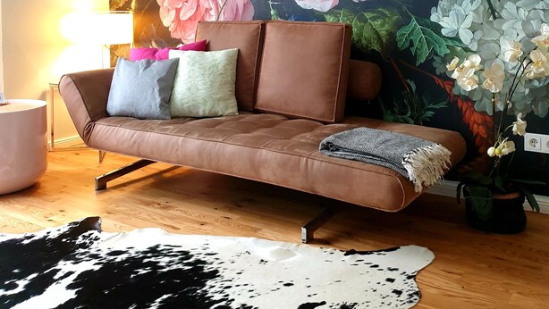 A living room with a brown leather sofa, a round coffee table, and a cowhide rug. A large floral mural is on the wall behind the sofa. - Apartment P1 in Berlin-Friedrichshain (Berlin)