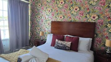 A hotel room with a floral wallpaper, a wooden headboard, a bed with white linens, two pillows, a bedside table with a lamp, and a window with curtains.