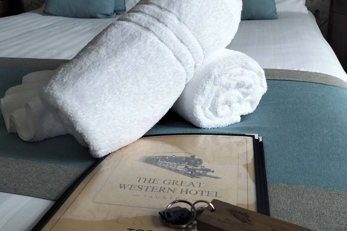 Ein ordentlich bezogenes Bett mit weißen Handtüchern, einer türkisblauen Decke und einem gerahmten Schild mit der Aufschrift The Great Western Hotel.