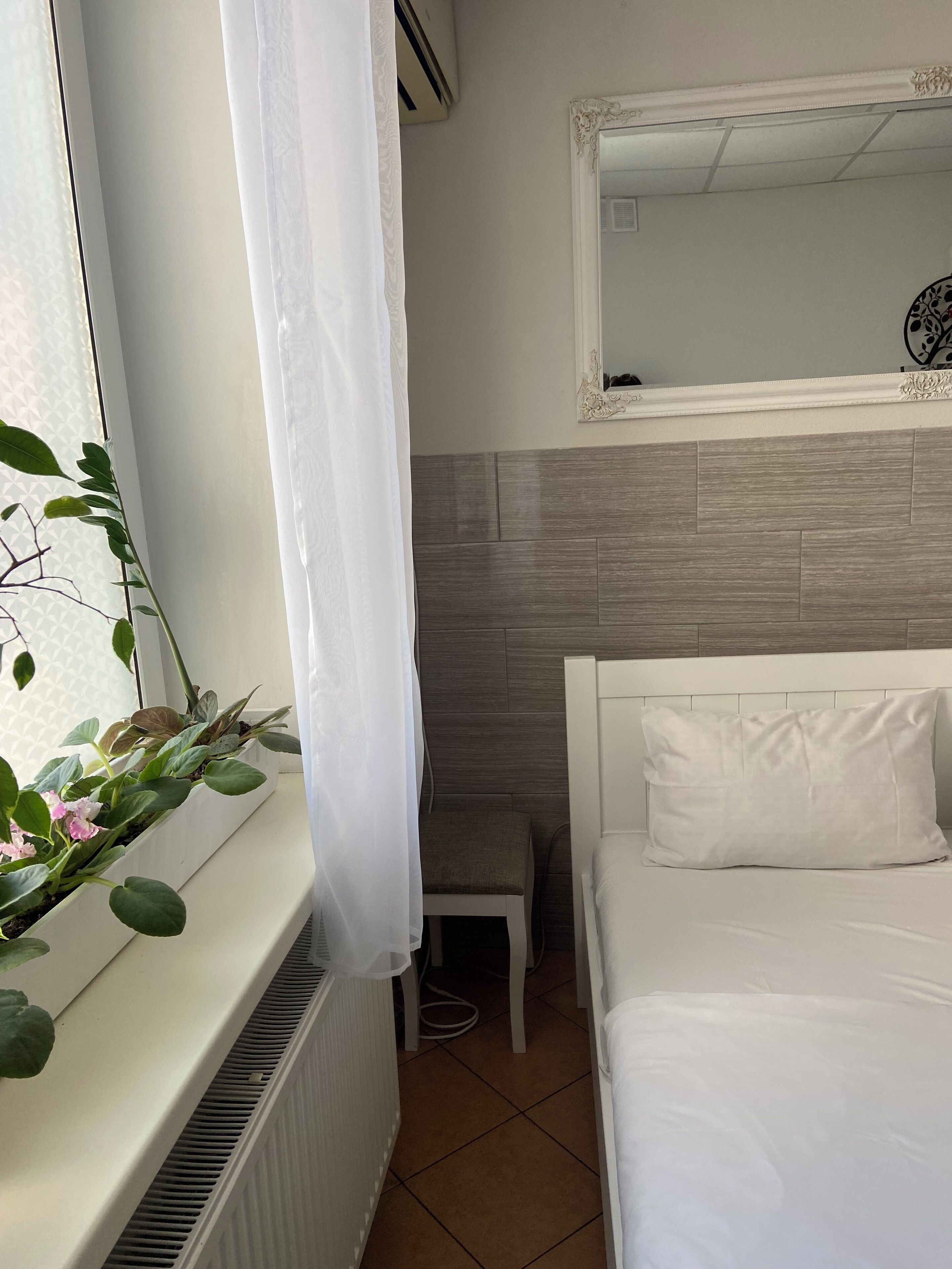 A bedroom with a white bed, a small bedside table, a radiator, a window with a curtain, and a mirror on the wall.