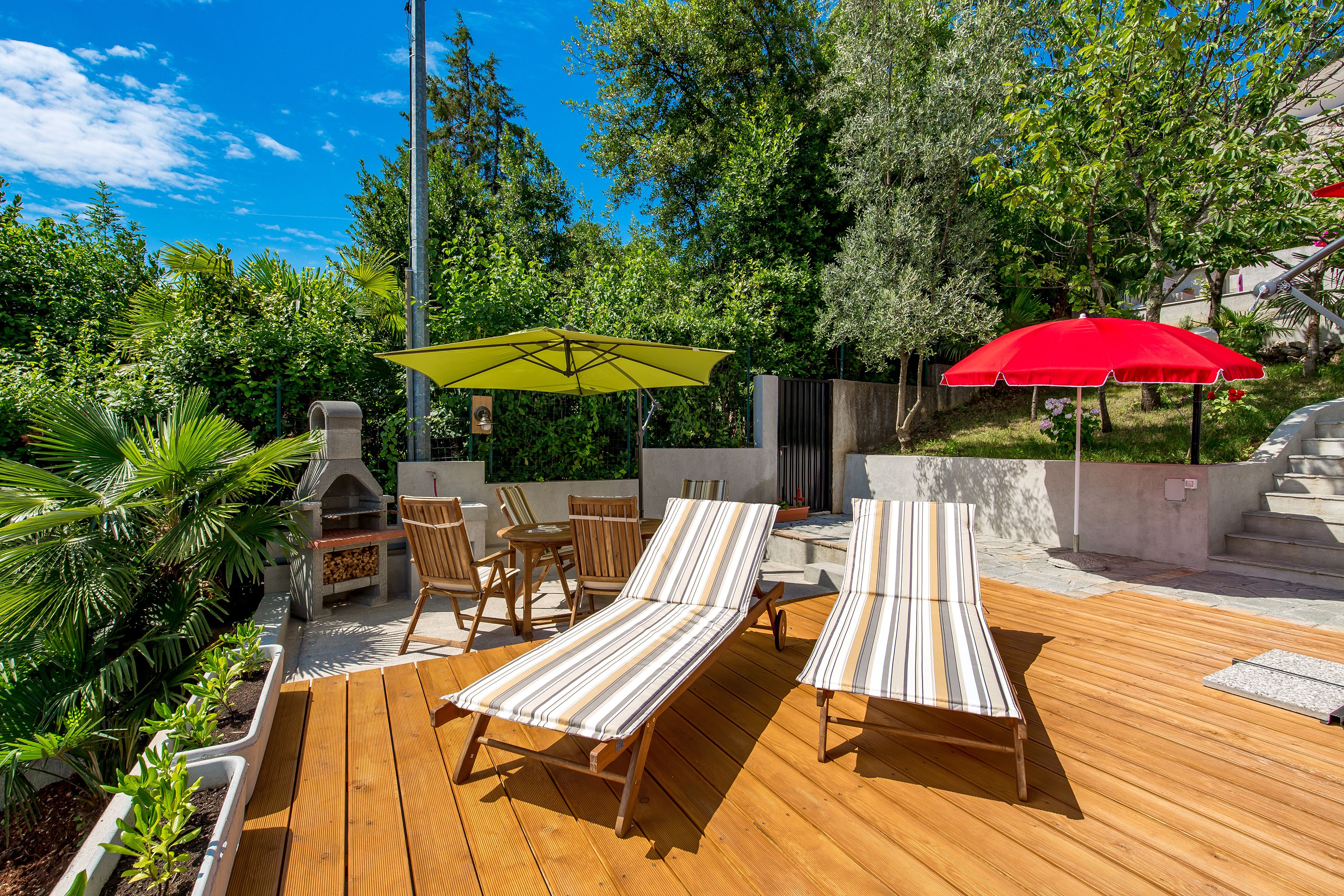 An outdoor patio with wooden flooring, striped lounge chairs, a barbecue grill, and two umbrellas.