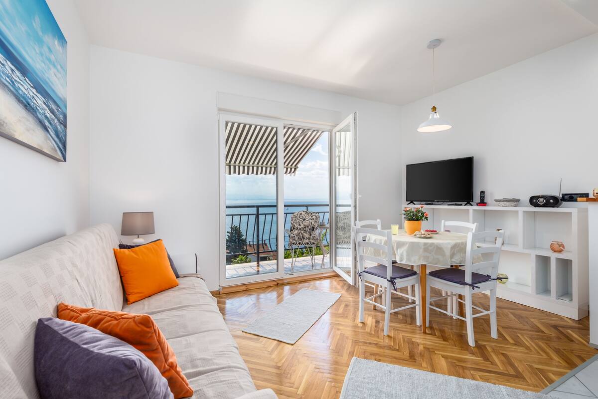 Un salon moderne avec un canapé, une table à manger et un téléviseur. Il y a un balcon avec vue sur la mer.