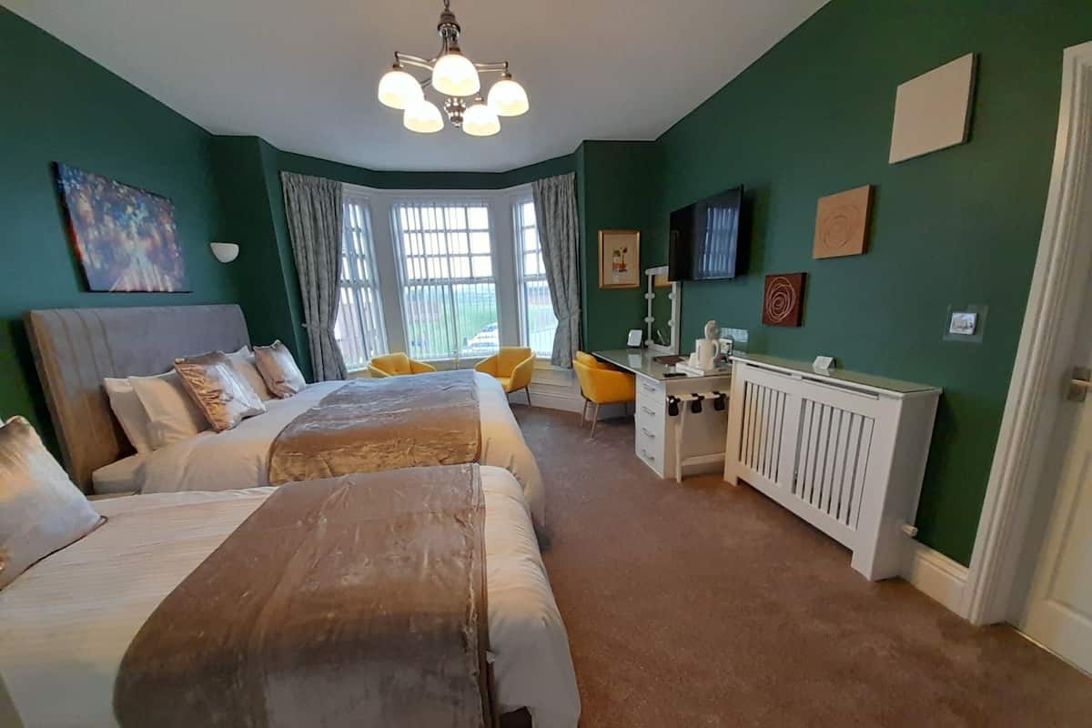 A bedroom with two beds, a desk, a radiator, and a large window with curtains.