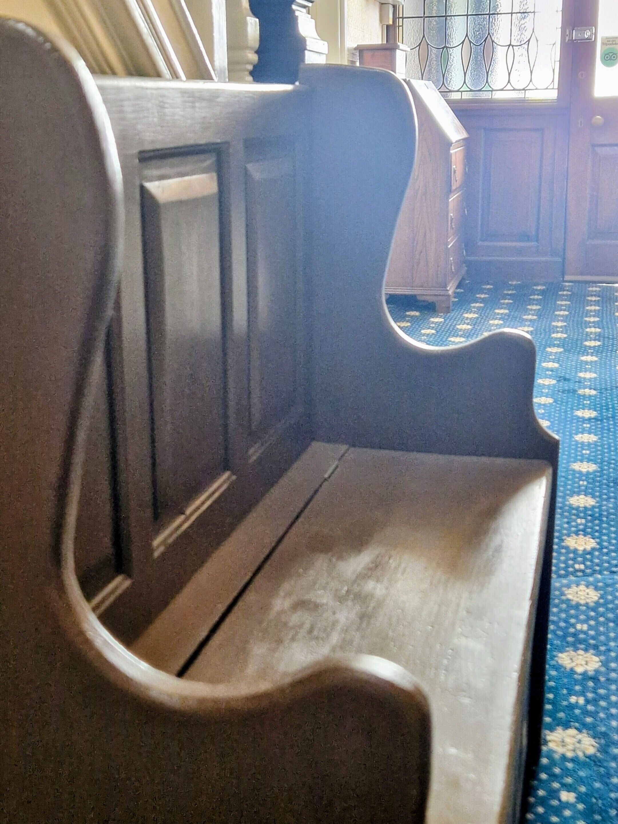 A wooden bench with a curved backrest and a flat seat, positioned in a room with patterned flooring and wooden furniture.