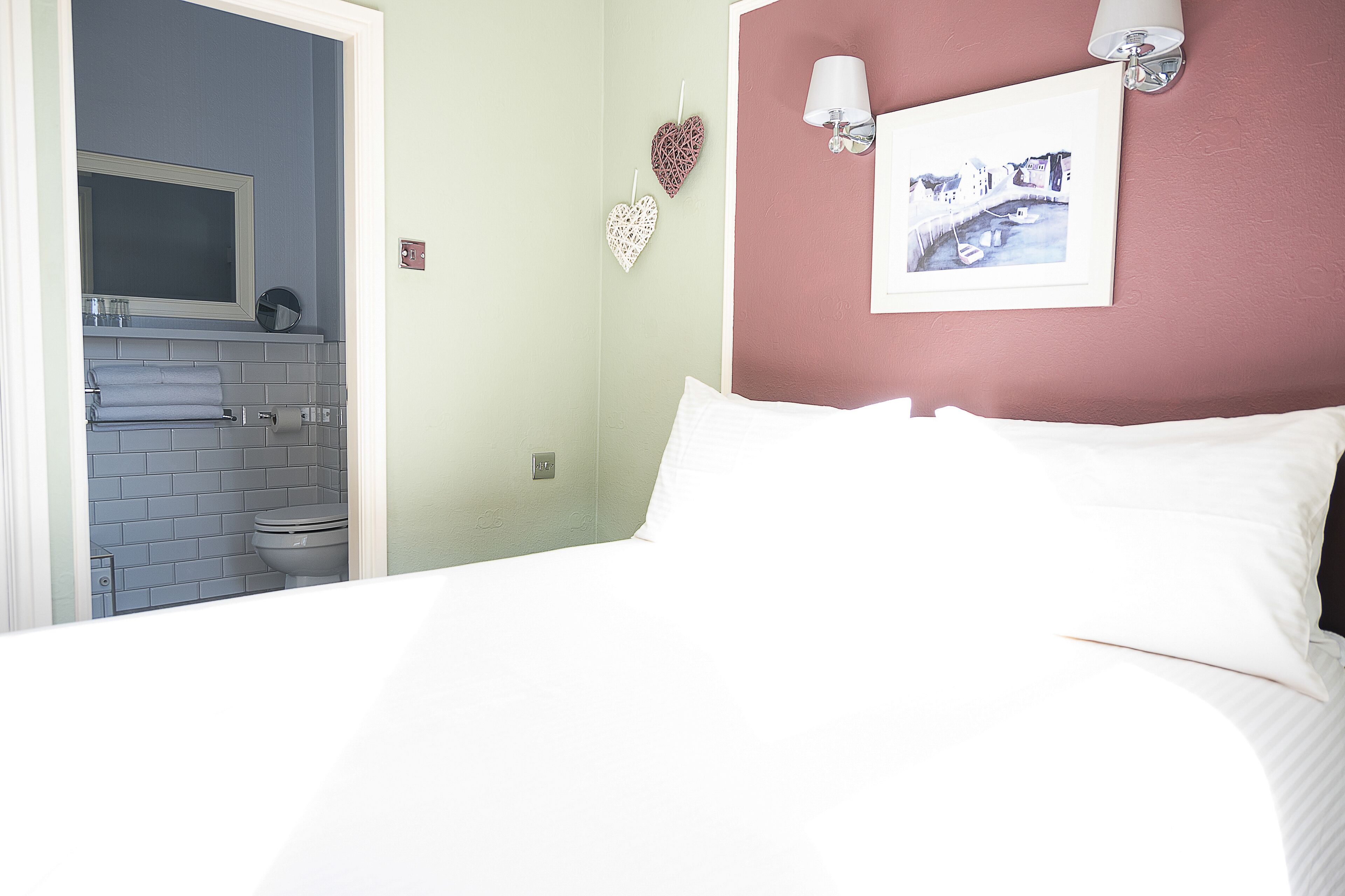 Ein Schlafzimmer mit Bett, einer weißen Wand mit einem gerahmten Bild und einem durch eine offene Tür einsehbaren Badezimmer.