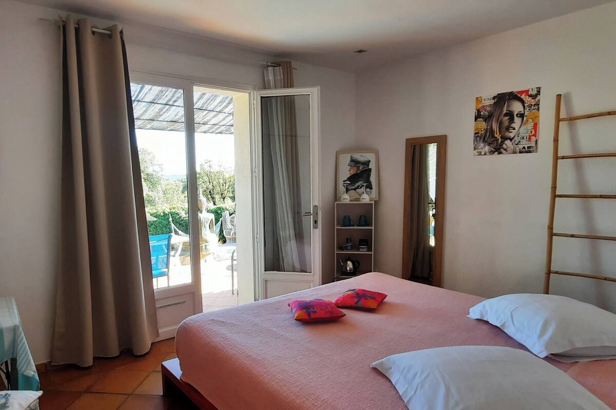 Un dormitorio con cama, escritorio y silla, televisor, ventana con cortinas y puerta que da a un área exterior.