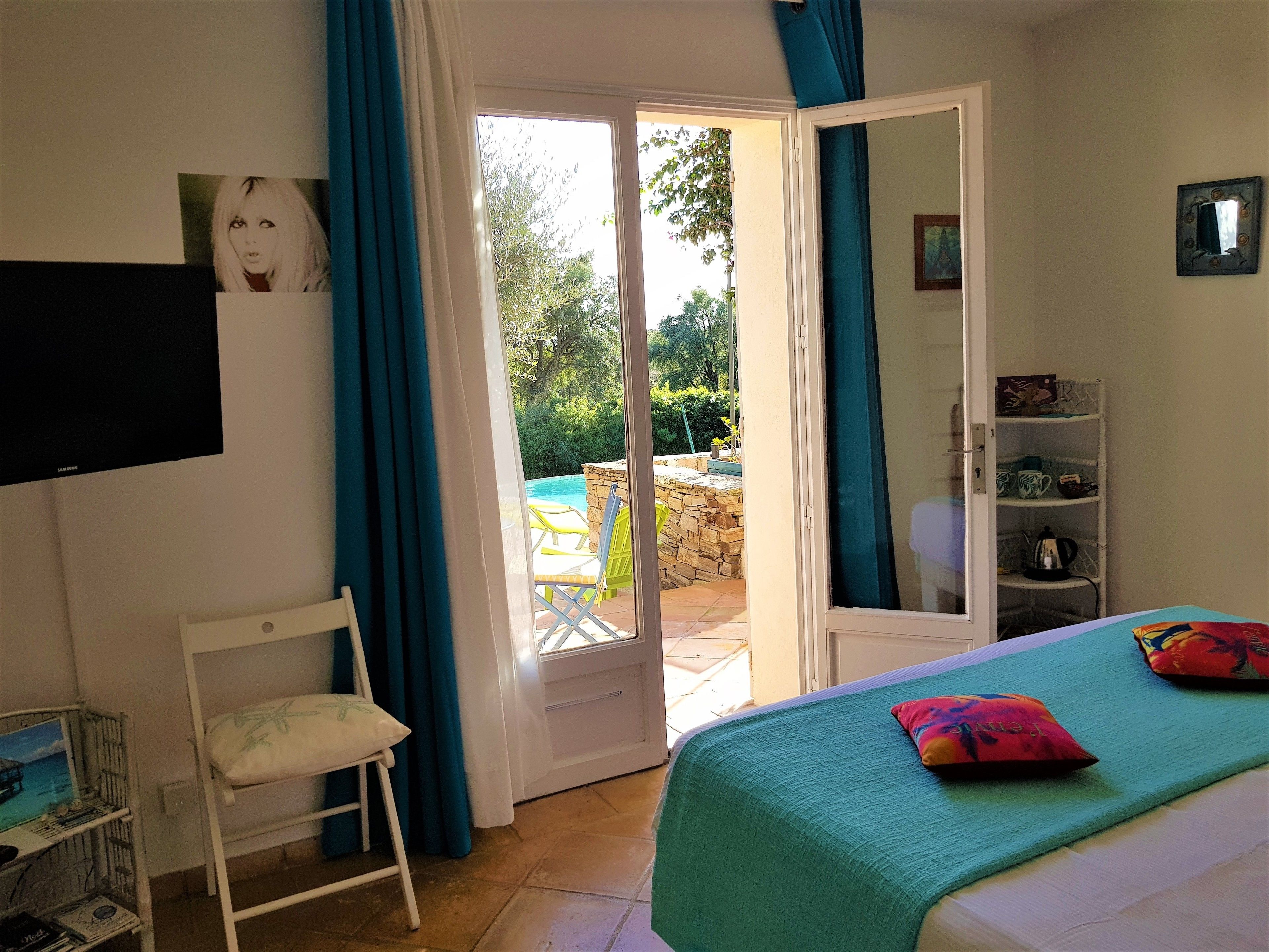 A bedroom with a bed, a chair, a TV, a shelf with items, and a door leading to a balcony with a pool.