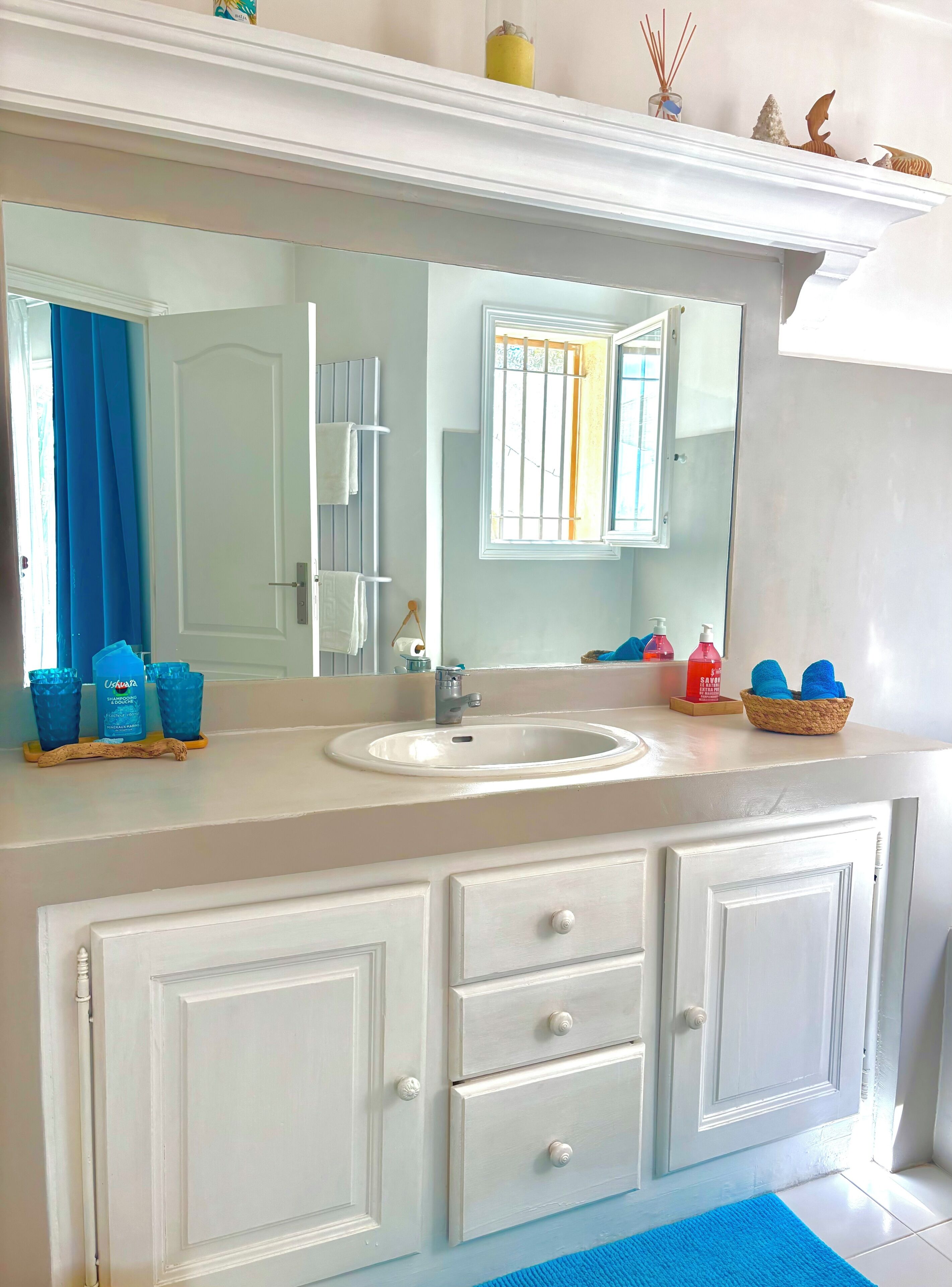 Une salle de bain avec un meuble-lavabo blanc, un grand miroir, un évier et un tapis de bain bleu.