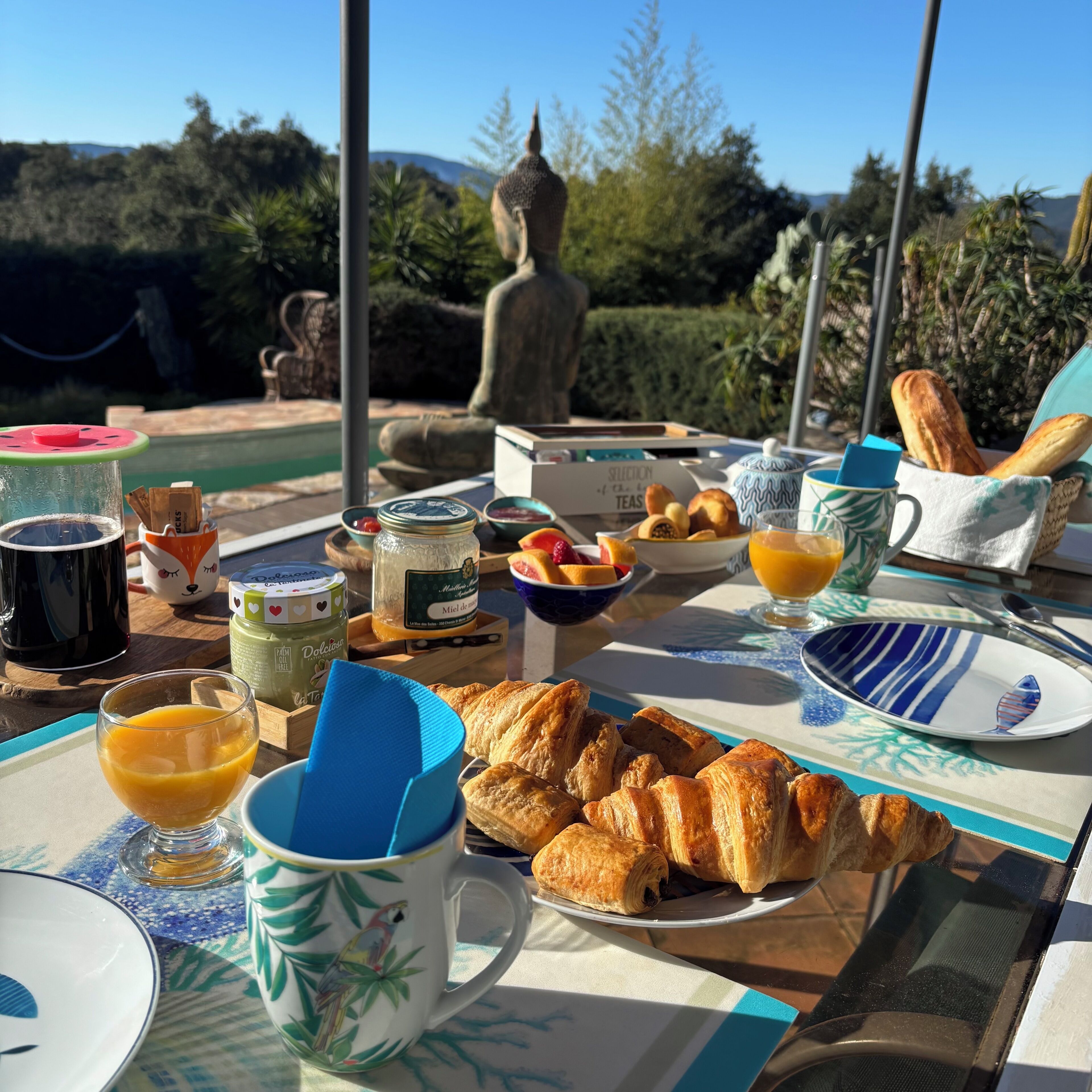 A breakfast table with croissants, fruit, and a Buddha statue in the background.