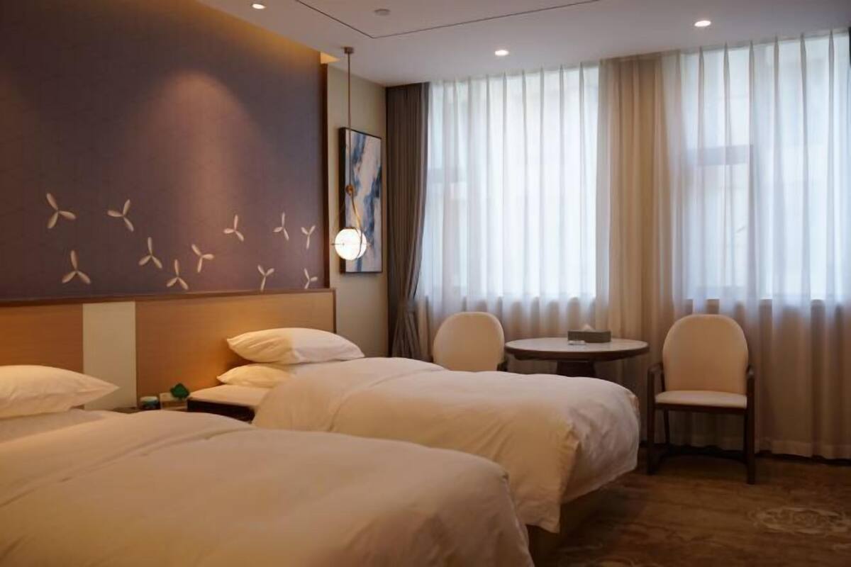 Camera d'albergo con due letti, un piccolo tavolo, una sedia e un'ampia finestra con tende.