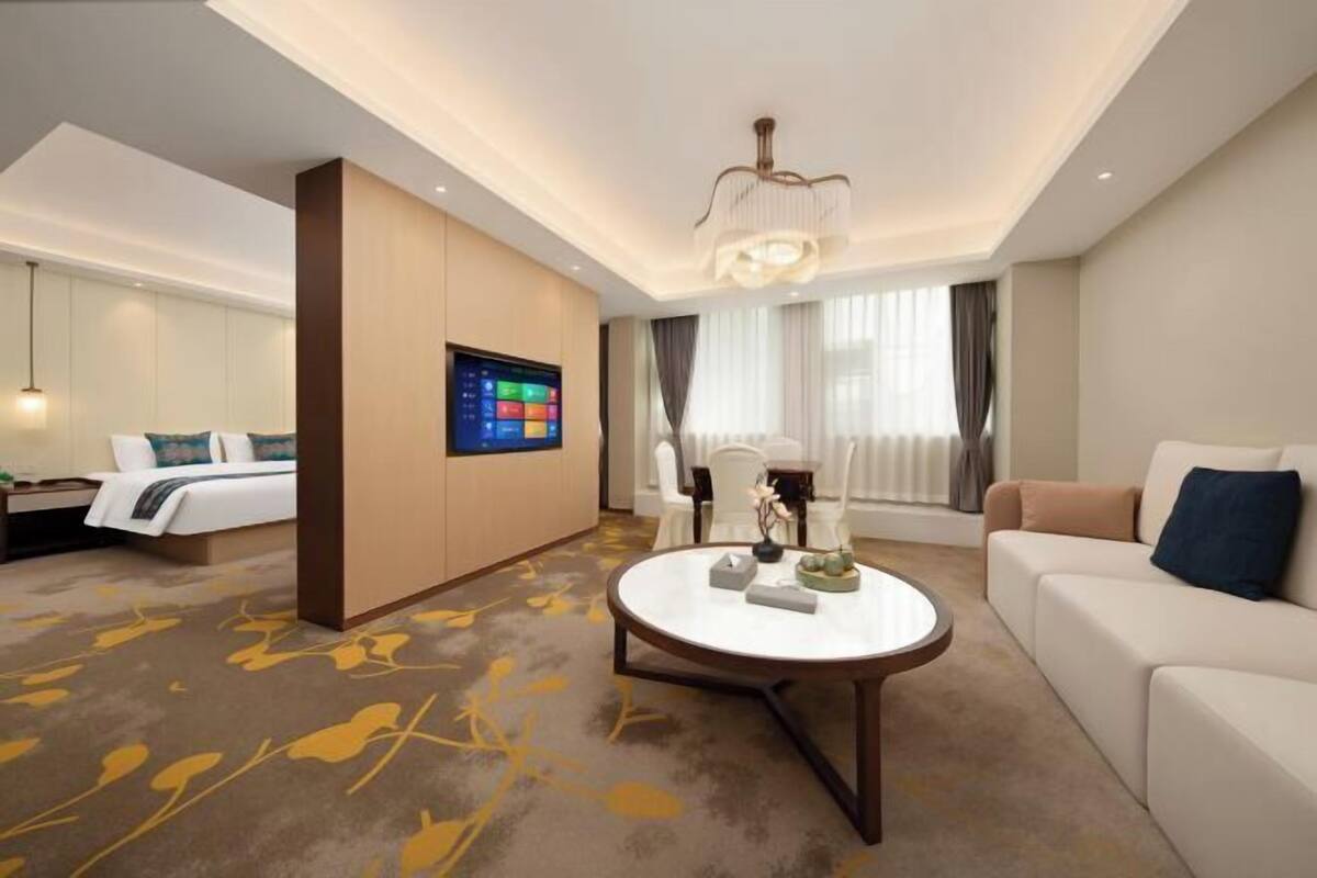 Una camera d'albergo con un letto, una TV, un tavolino rotondo, un divano e un'ampia finestra con tende.