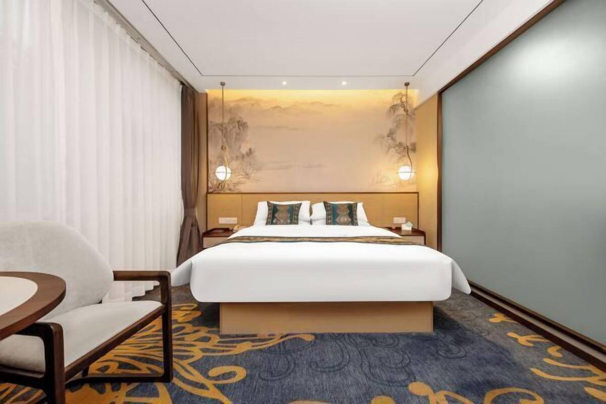 Una camera d'albergo con un letto grande, una sedia, un comodino e una parete con un dipinto di un paesaggio.