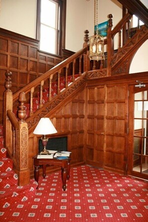 Una escalera de madera con intrincados adornos, un suelo de alfombra roja con diseño y una mesa con una lámpara y papeles.