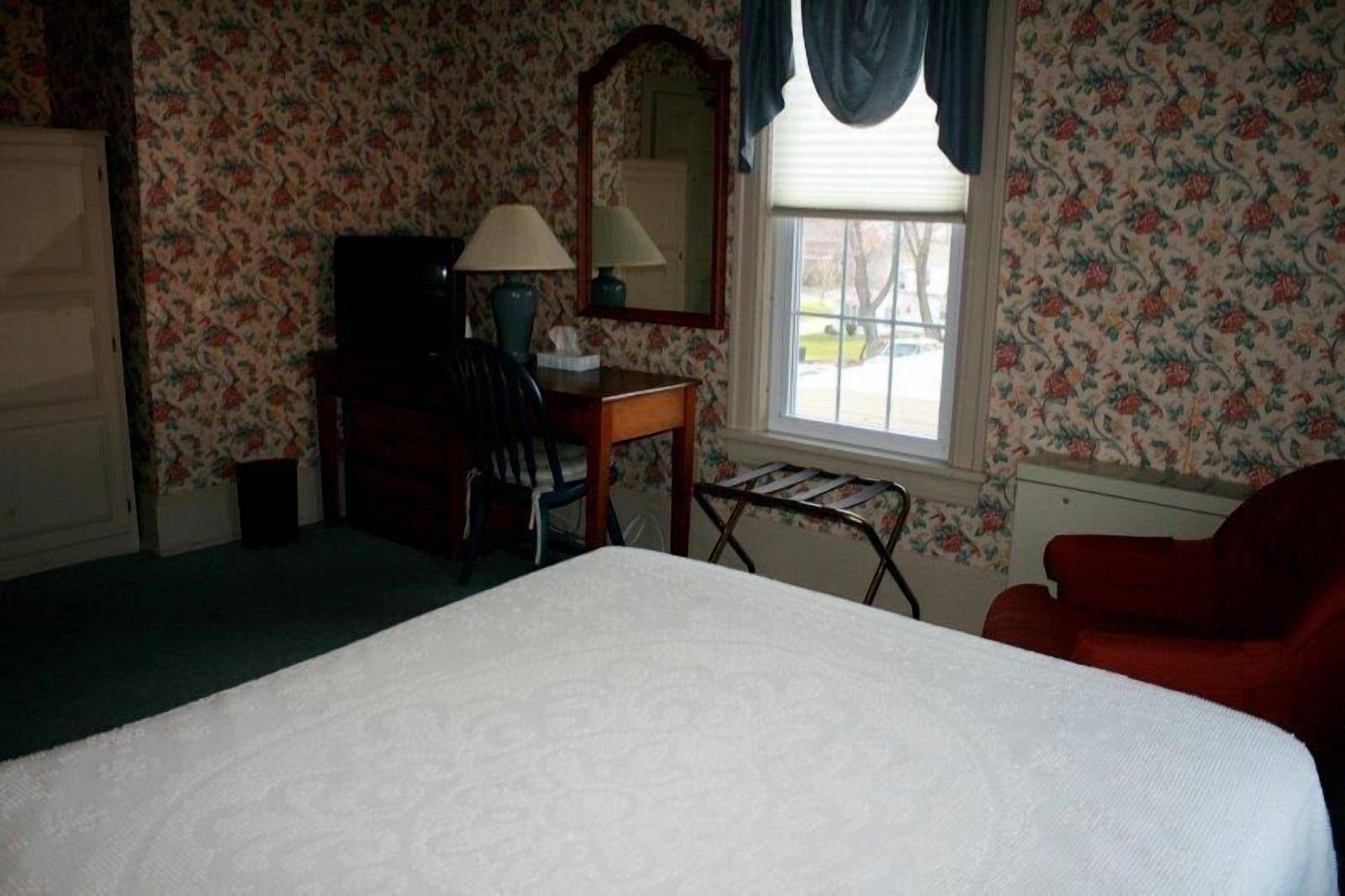A hotel room with floral wallpaper, a bed, a desk with a lamp, a chair, and a window with curtains.
