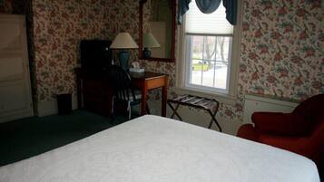 A hotel room with floral wallpaper, a bed, a desk with a lamp, a chair, and a window with curtains.