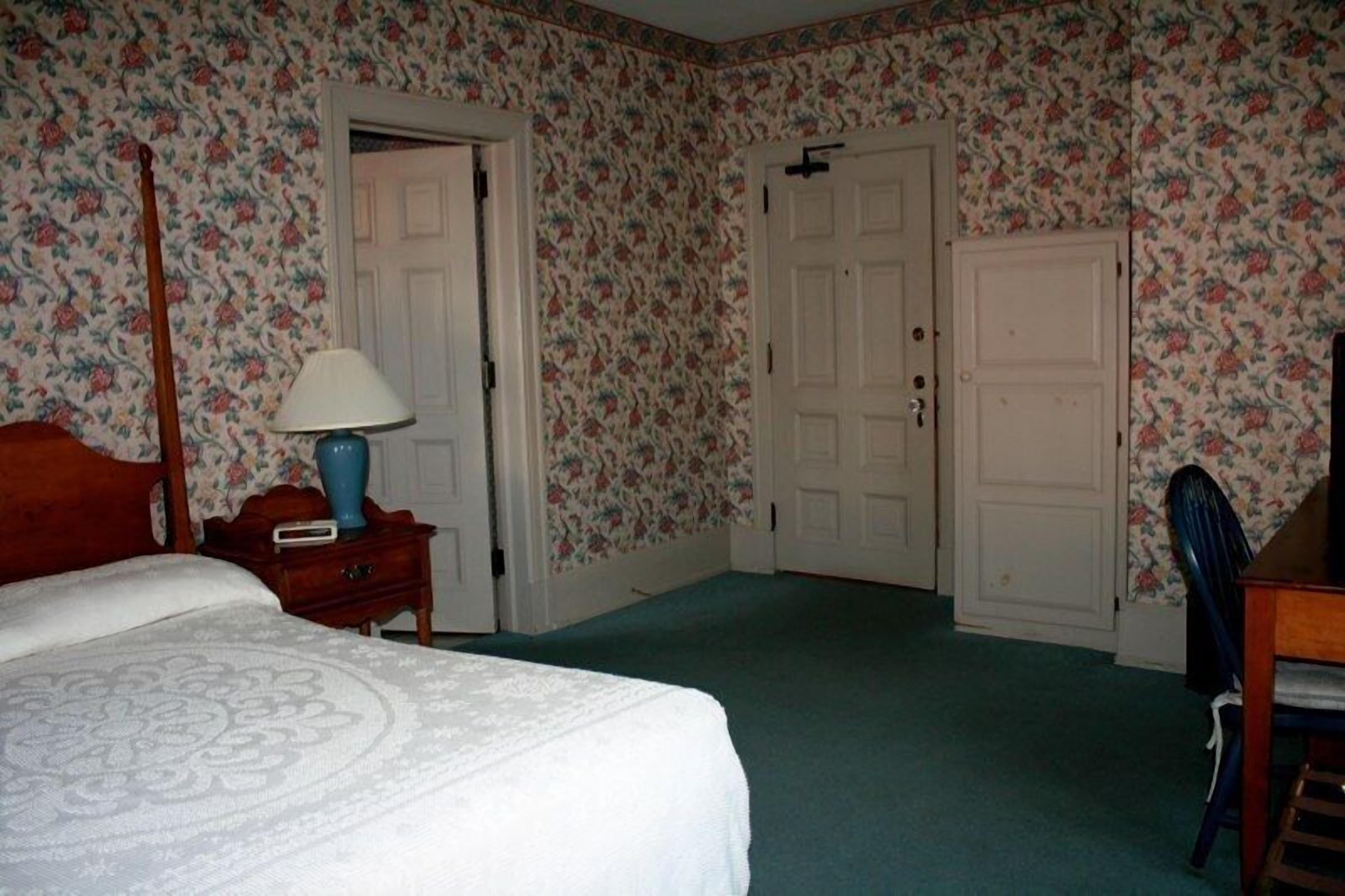 Un dormitorio con papel tapiz floral, una cama con cubrecama blanco, una mesita de noche de madera con lámpara, y una silla.