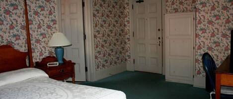 Un dormitorio con papel tapiz floral, una cama con cubrecama blanco, una mesita de noche de madera con lámpara, y una silla.