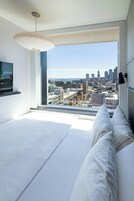 A modern bedroom with a large window offering a city view, a bed with white linens, and a mounted TV on the wall.