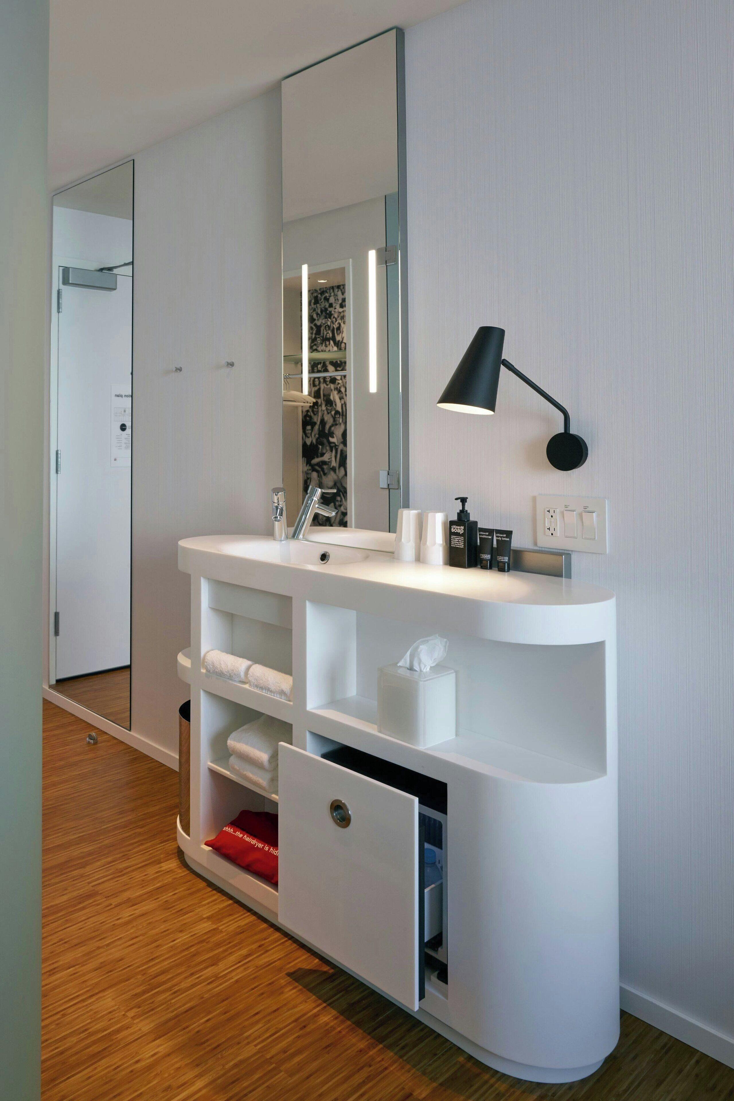 A modern bathroom with a white vanity, a large mirror, a black wall-mounted lamp, and a wooden floor.