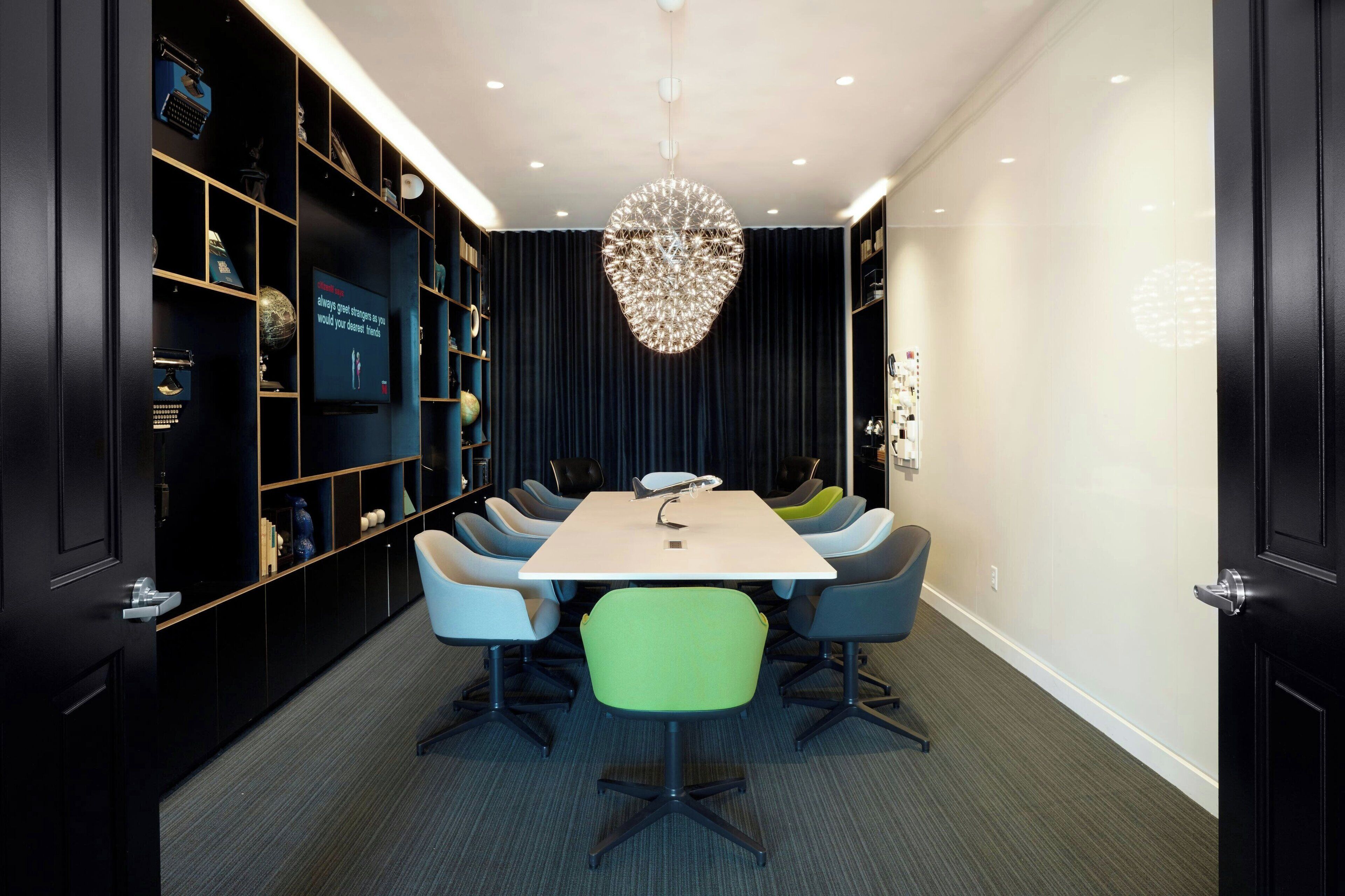 A conference room with a white table, blue and green chairs, a chandelier, and black shelving with various items.