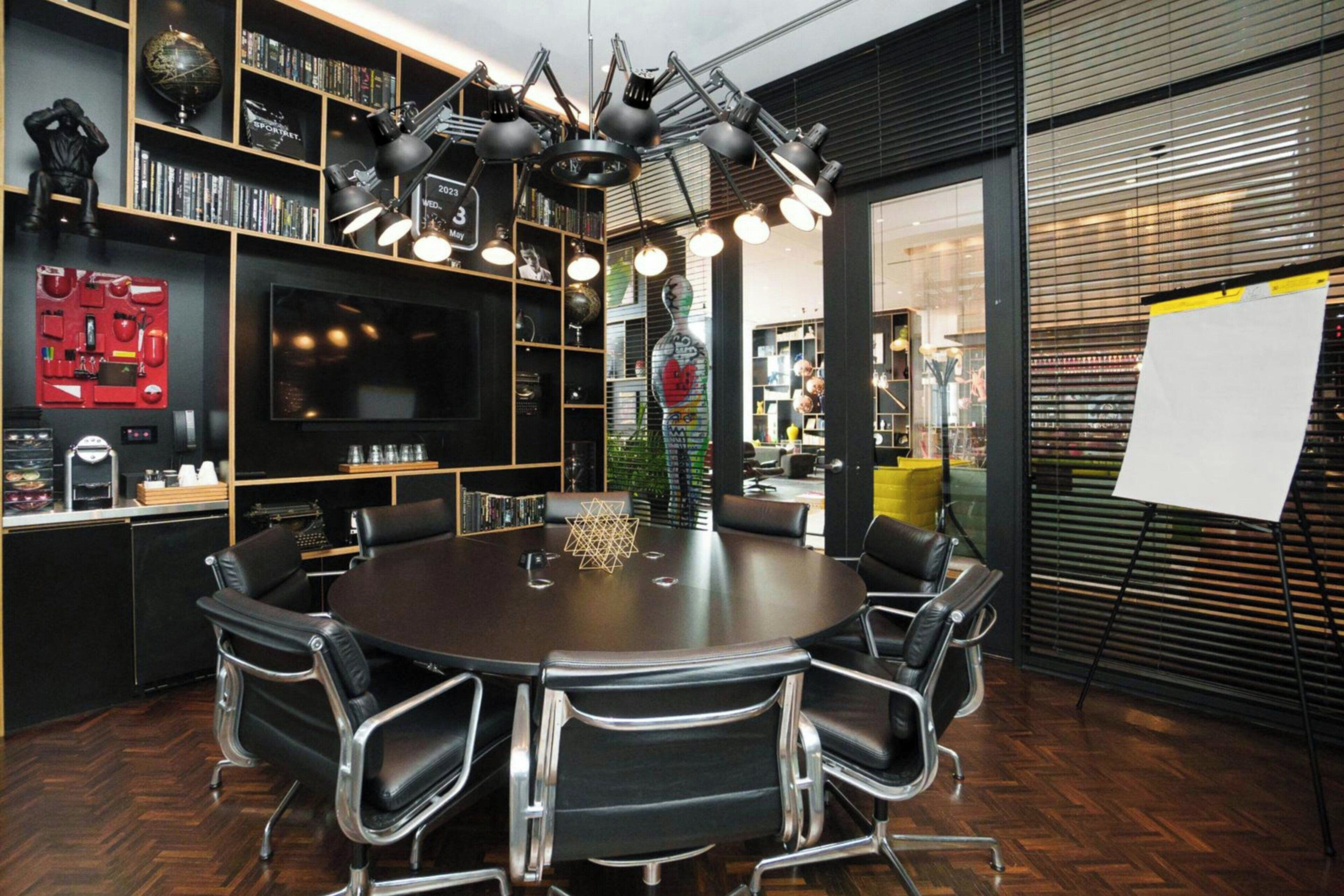 Une salle de conférence équipée d’une table ronde, de fauteuils en cuir noir, d ’un grand téléviseur et d’étagères remplies de livres et d’objets décoratifs.