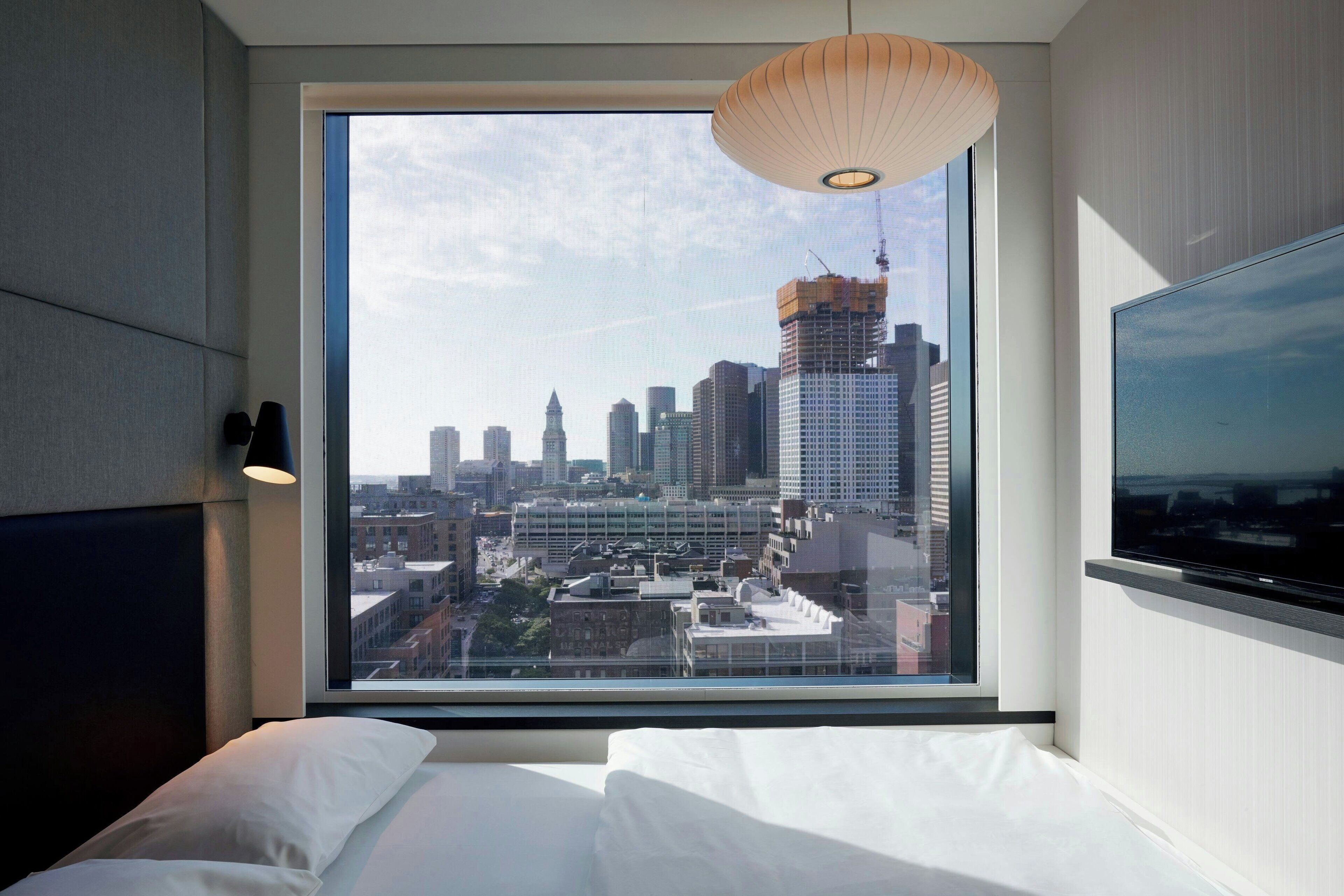 Camera d'albergo con un'ampia finestra che si affaccia sulla città, un letto con lenzuola bianche e una moderna lampada a sospensione.