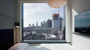 Camera d'albergo con un'ampia finestra che si affaccia sulla città, un letto con lenzuola bianche e una moderna lampada a sospensione.
