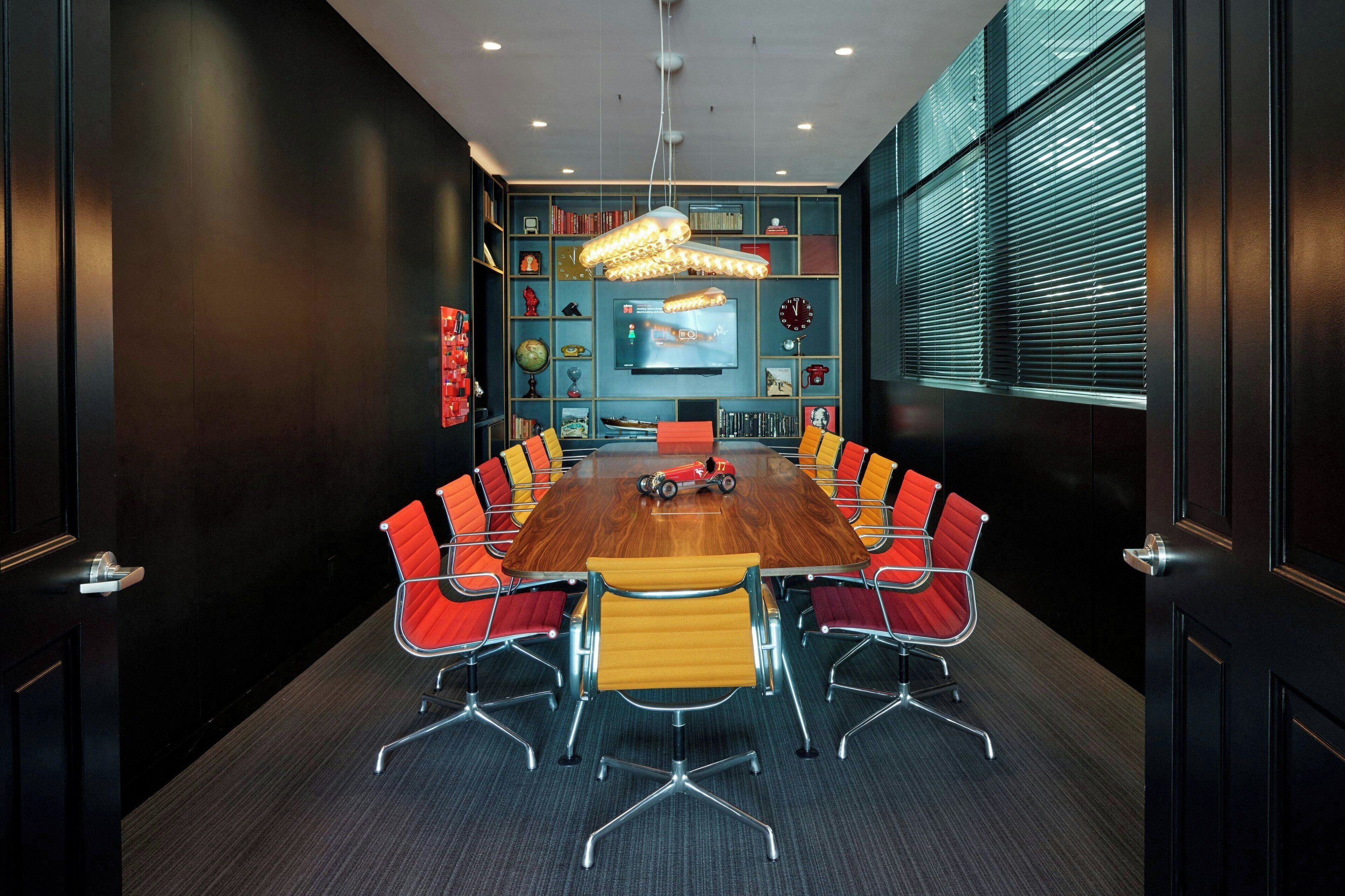 A conference room with a wooden table, red and yellow chairs, a television, and a bookshelf with various items.