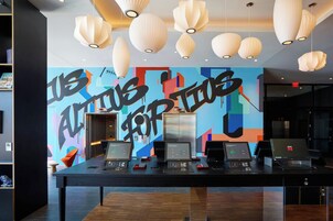 Un'area di lavoro moderna con diversi portatili su un tavolo lungo, un murale astratto dai colori vivaci al muro e luci a sospensione.