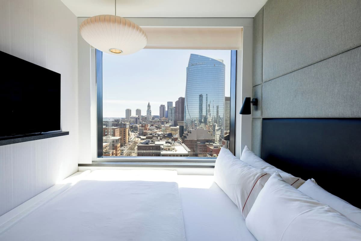 Ein modernes Hotelzimmer mit einem großen Bett, einem an der Wand befestigten Flachbildfernseher und Blick über die Stadt durch ein großes Fenster.