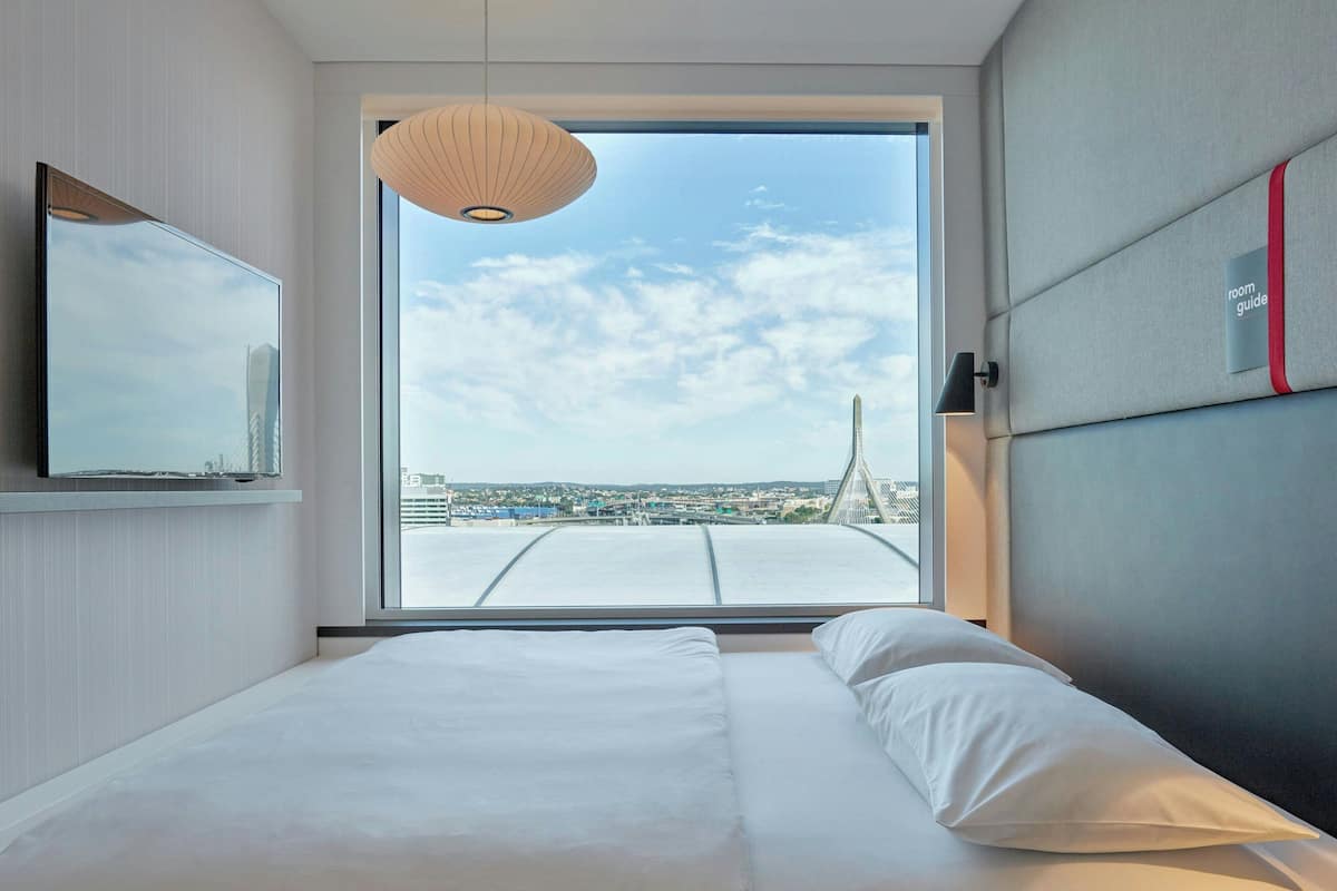 Ein modernes Hotelzimmer mit einem großen Bett, einem Fernseher und Blick auf eine Stadtlandschaft durch ein großes Fenster.