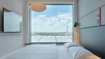 A modern hotel room with a large bed, a television, and a view of a cityscape through a large window.
