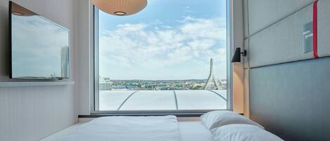 Quarto de hotel moderno com uma cama grande, televisão e vista para o horizonte urbano através de uma janela ampla.