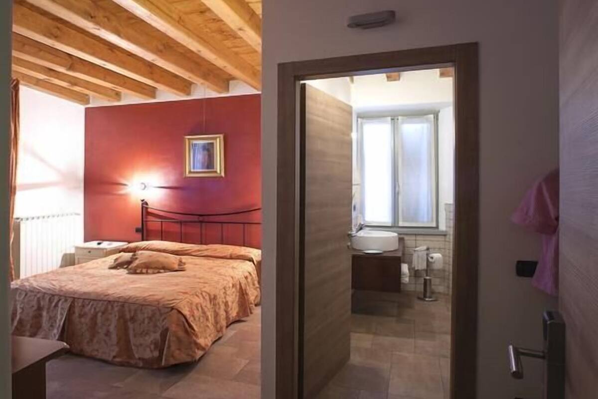 Ein Schlafzimmer mit Bett, Nachttisch und roter Wand. Die Decke ist aus Holz und eine Tür führt ins Badezimmer.