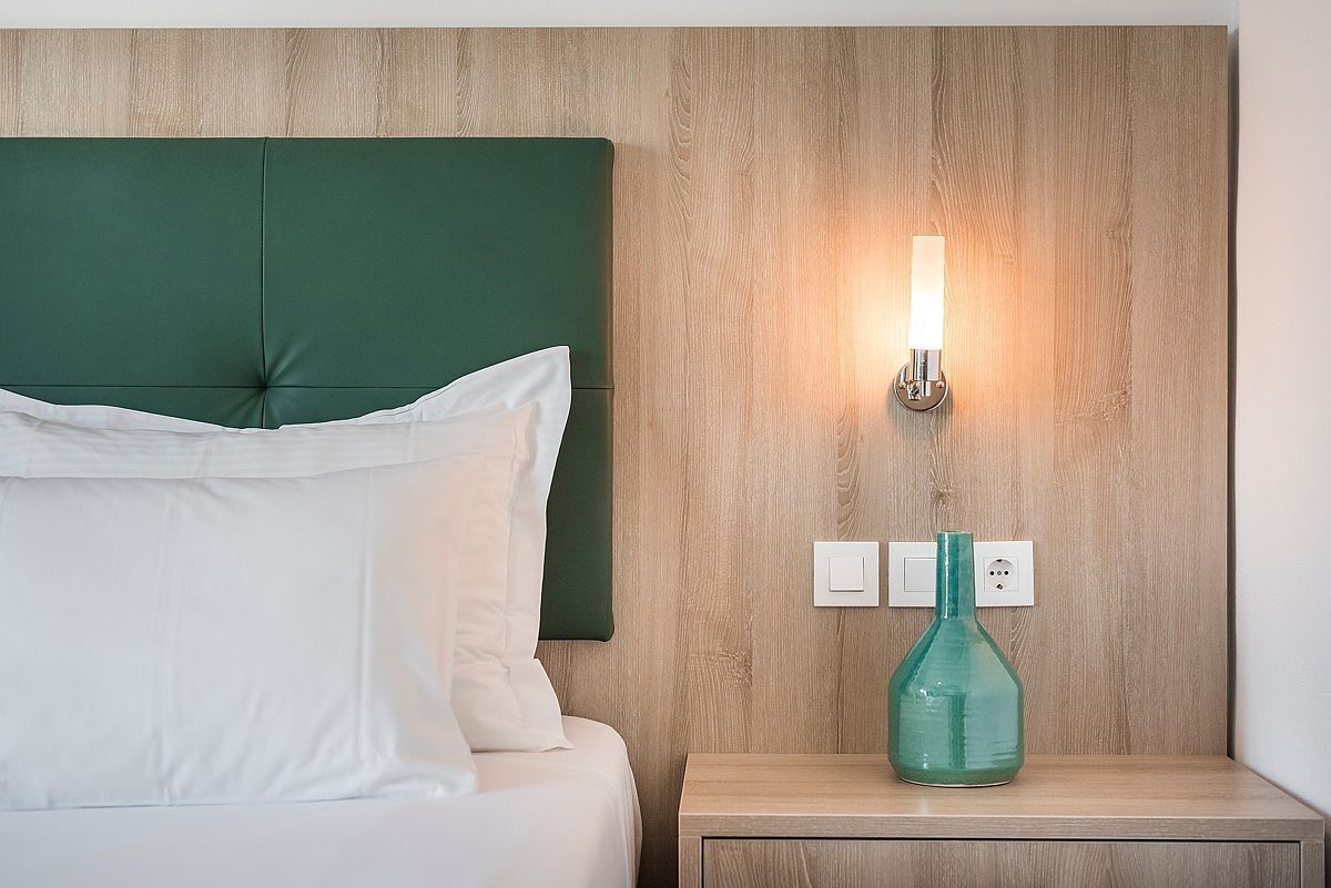 Una cama bien hecha con sábanas blancas y un cabecero verde, una mesita de noche con un jarrón verde, y una lámpara de pared.