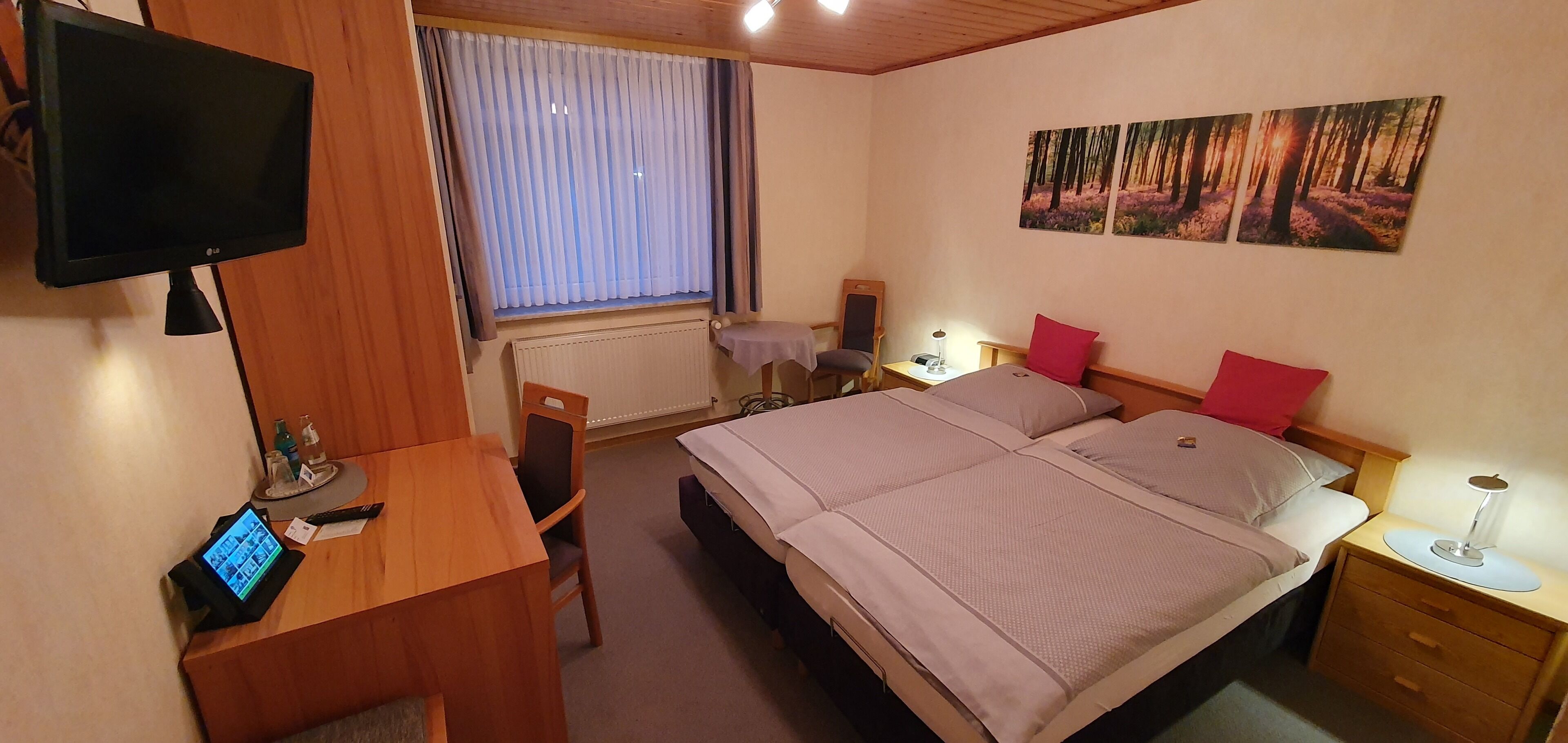 A hotel room with a bed, bedside table, lamp, desk with a computer, chair, and a TV.