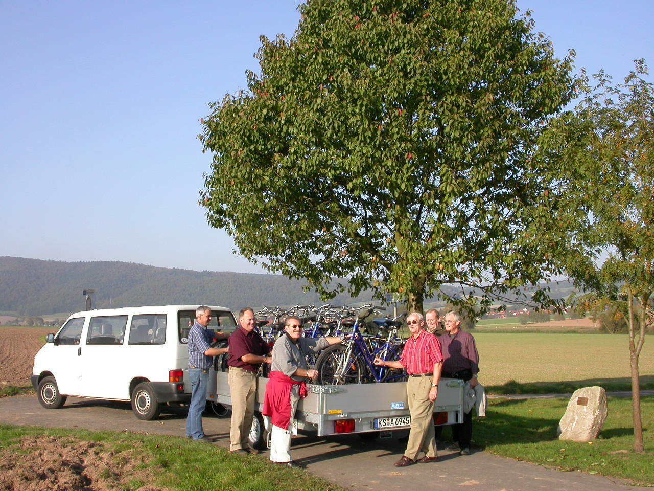 A group of people standing by a white van with bicycles mounted on the back, parked on a rural road with a tree and open fields in the background.