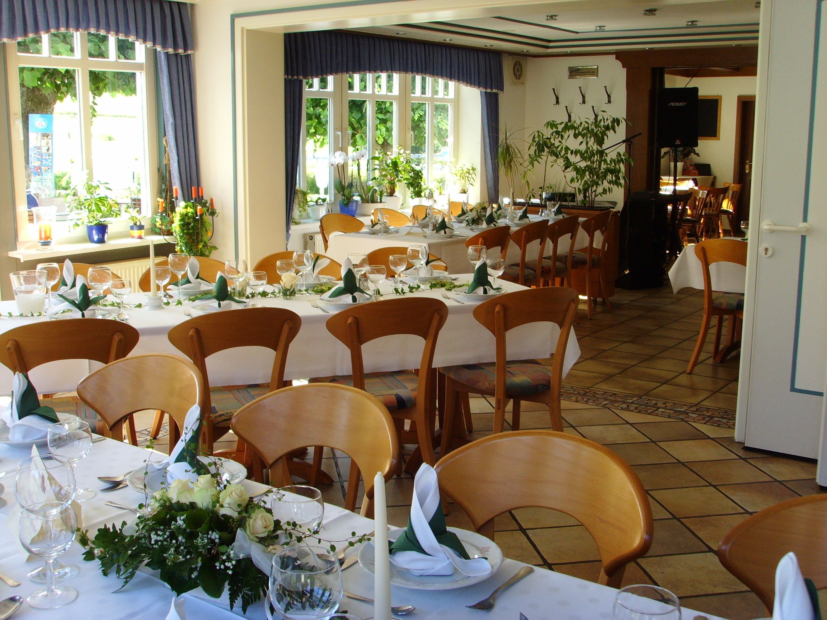 A restaurant with multiple tables set for dining, wooden chairs, and potted plants.
