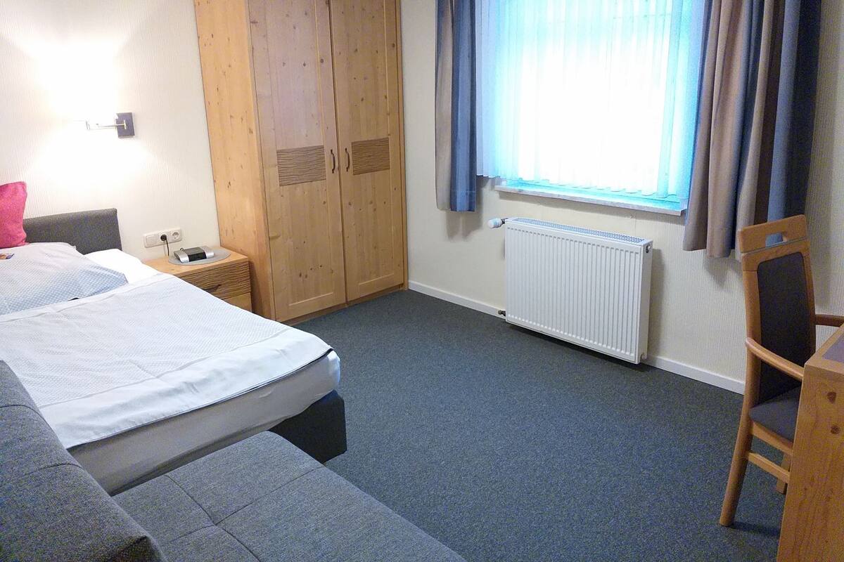 Une chambre d’hôtel avec un lit, une armoire, un radiateur et une fenêtre avec des rideaux.