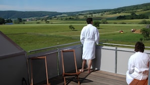 Een balkon met twee houten stoelen en een houten bankje, met uitzicht op een groen landschap met velden en heuvels in de verte.