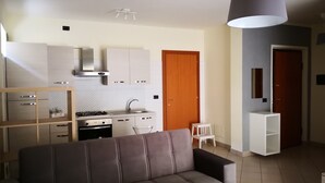 A modern living room with a grey sofa, a kitchen area with white cabinets, a stove, and a wooden door.