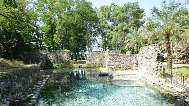 A clear pool surrounded by stone walls and lush greenery, with a stone bridge in the background.