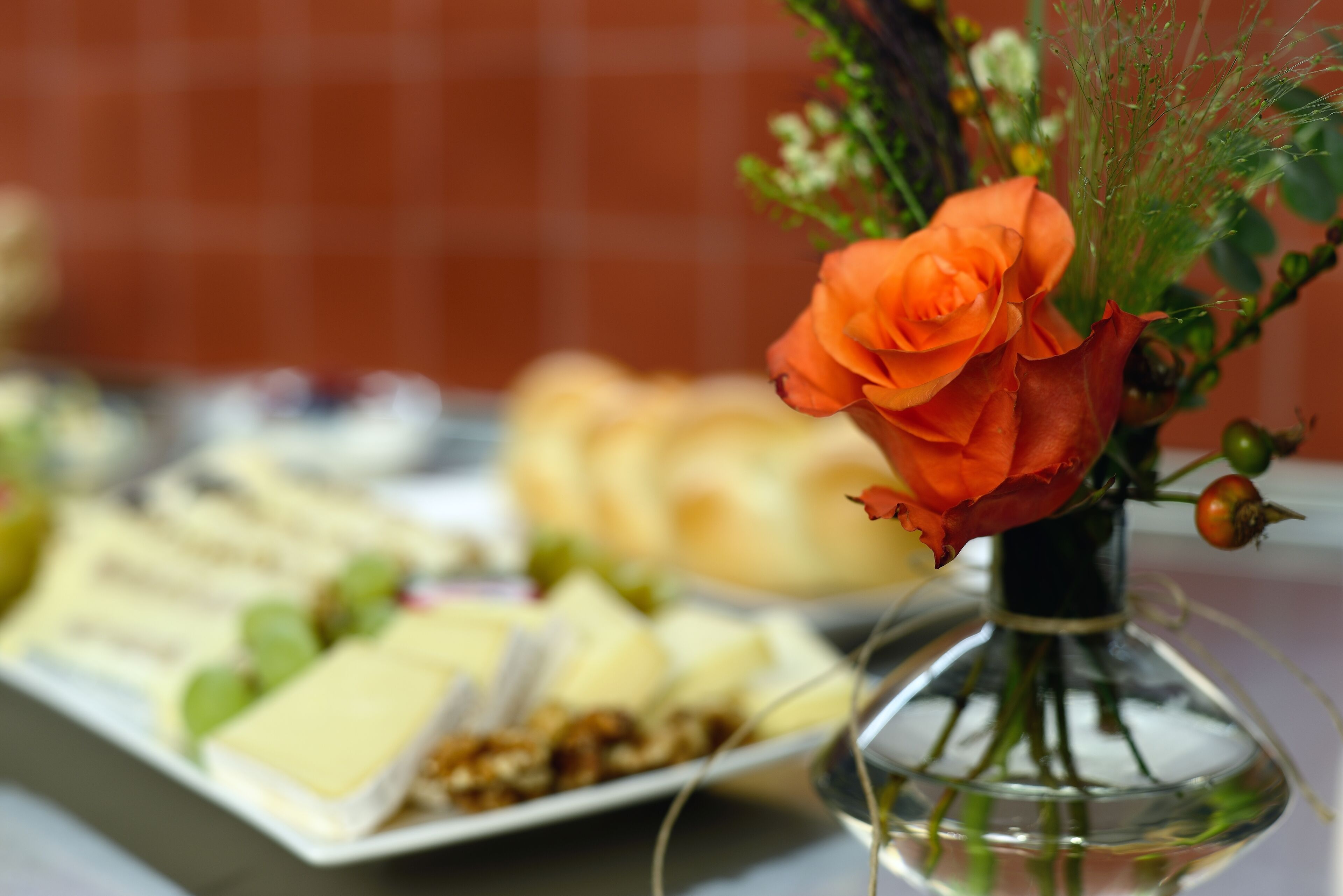 A table with a glass vase containing a single orange rose and some greenery, alongside a plate of assorted cheeses and crackers.