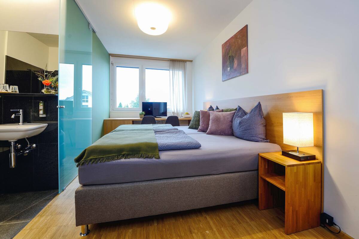 A modern hotel room with a bed, bedside table, lamp, and a view of the outside through large windows.